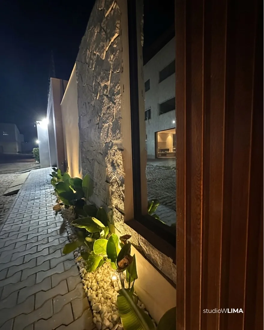 Lateral da Casa Comemorar à noite, com parede de pedra, jardim iluminado e piso intertravado