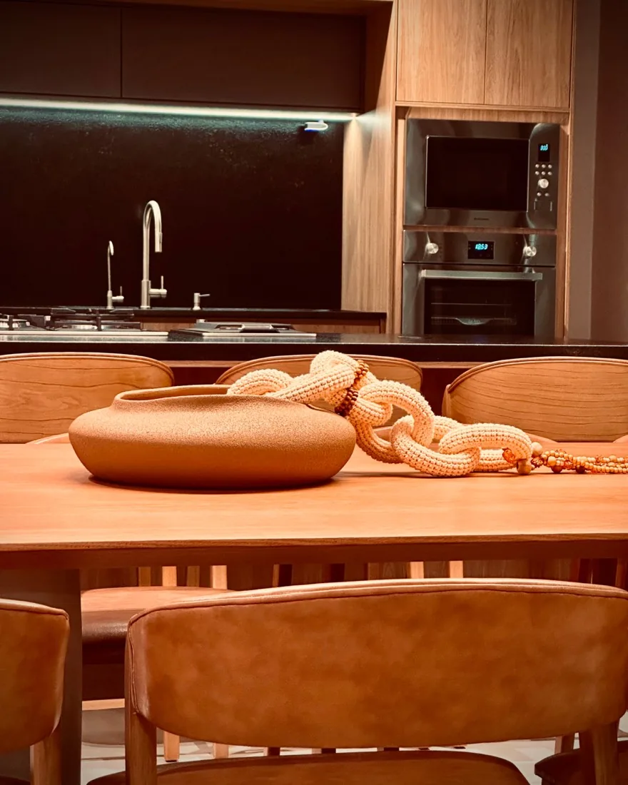Mesa de jantar em madeira com cadeiras estofadas e cozinha gourmet ao fundo na Casa Comemorar