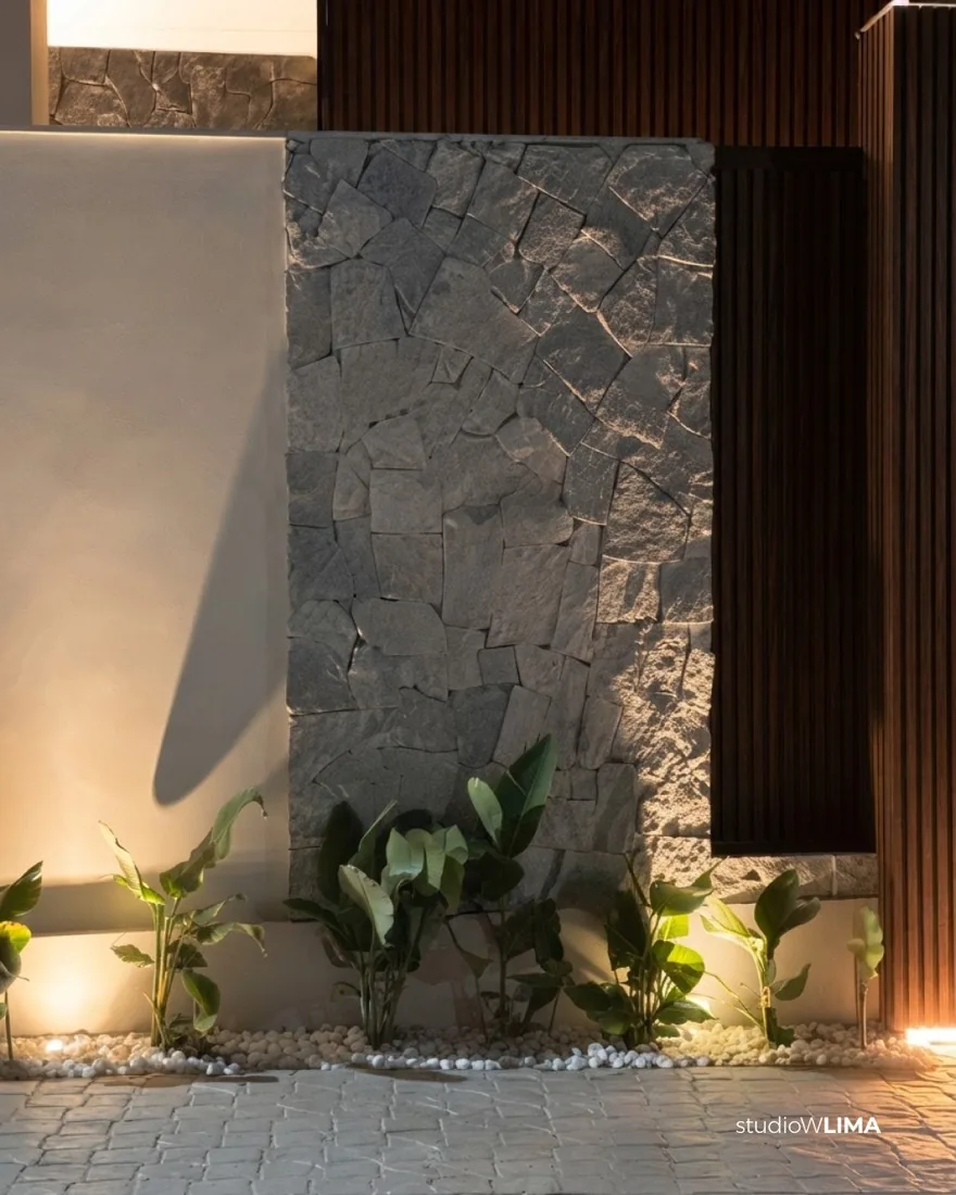 Muro de pedra natural cinza com iluminação de jardim e plantas na entrada da Casa Comemorar