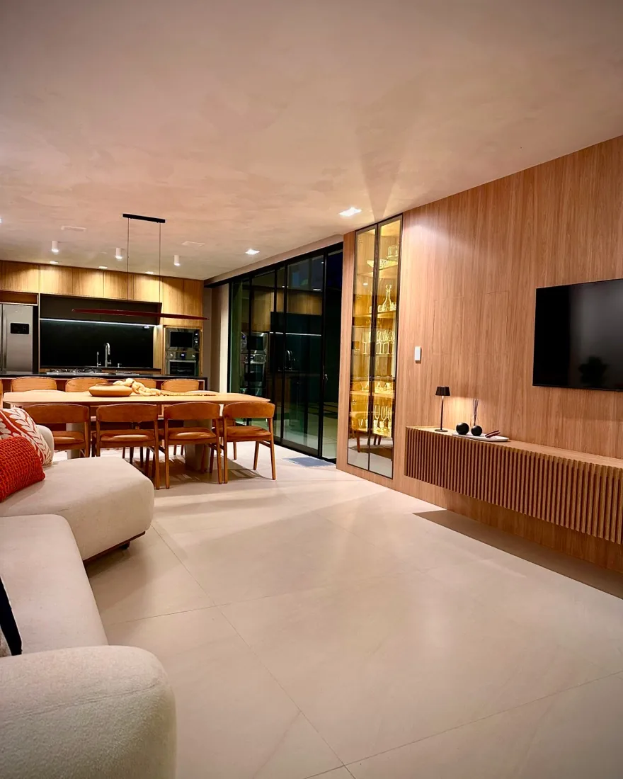 Sala de estar e jantar integradas da Casa Comemorar, com painel e rack em madeira ripada, sofá curvo claro e cozinha ao fundo