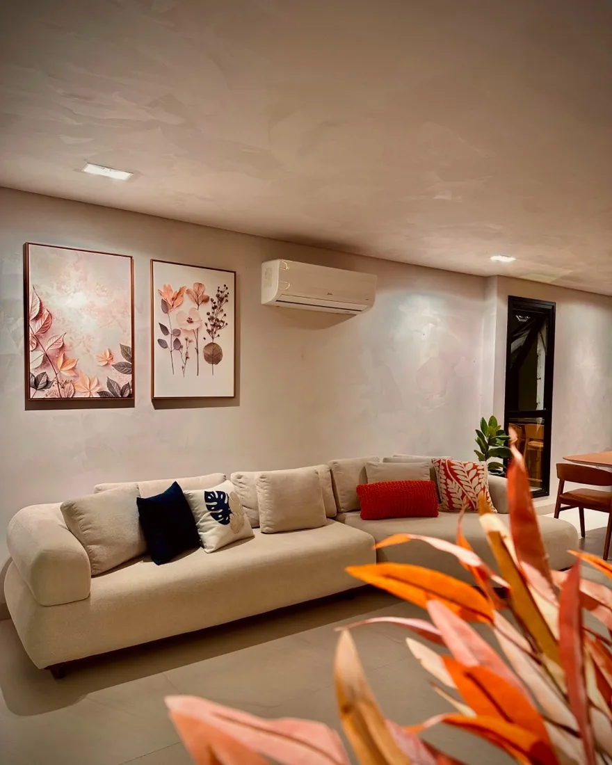 Sala de estar da Casa Comemorar com sofá bege, almofadas coloridas, quadros botânicos e paredes em acabamento claro